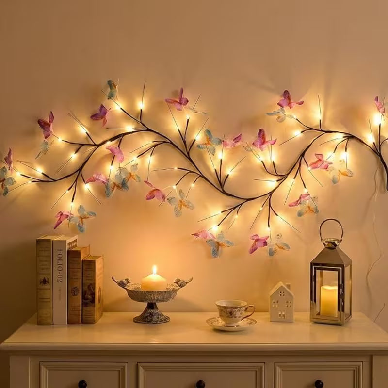 Branch-shaped fairy string lights with colorful butterfly decorations mounted on a beige wall above a console table with books, a candle, a teacup, and a lantern.